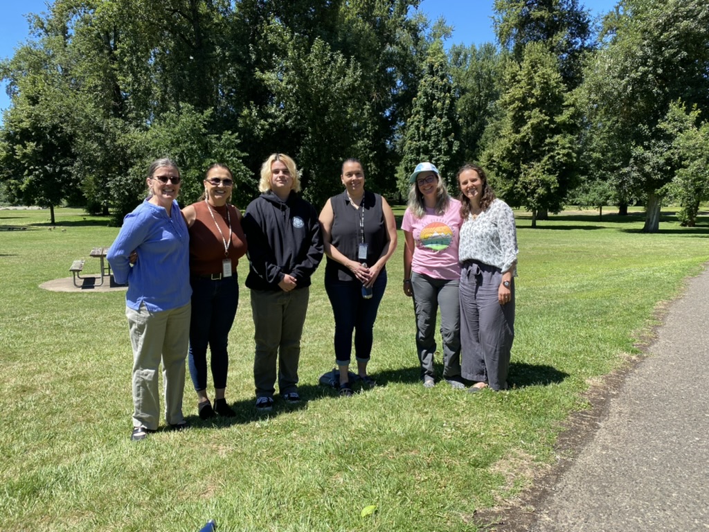 A group of six people standing together in a semi-circle. They are in a park-like setting with green grass, trees, and a picnic table behind them. This group includes unnamed Neuro staff and Spirit Mountain Community Fund staff - all smiling.