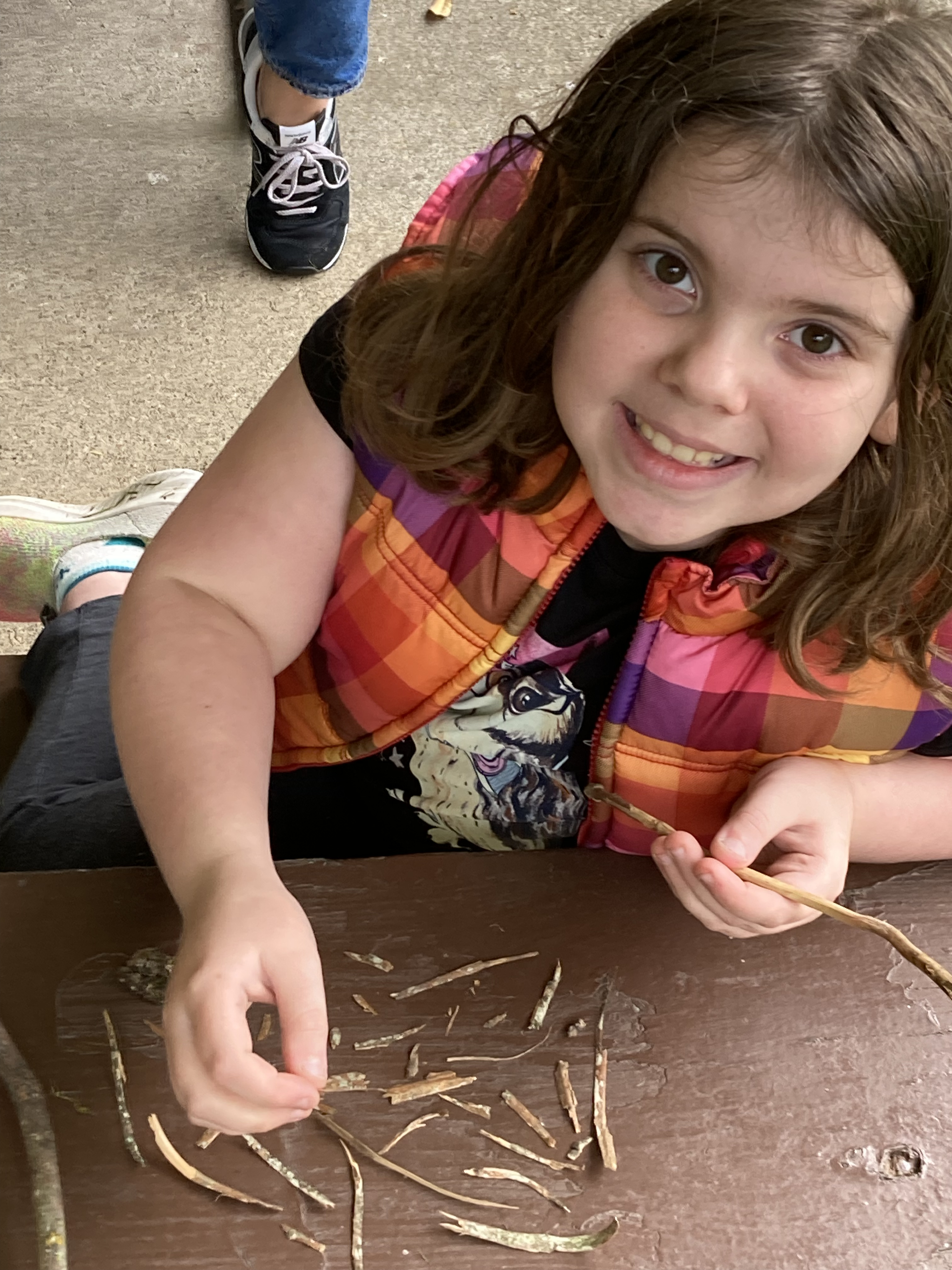 A little girl smiling at the camera as she plays with small tree branches for her nature based therapy session.