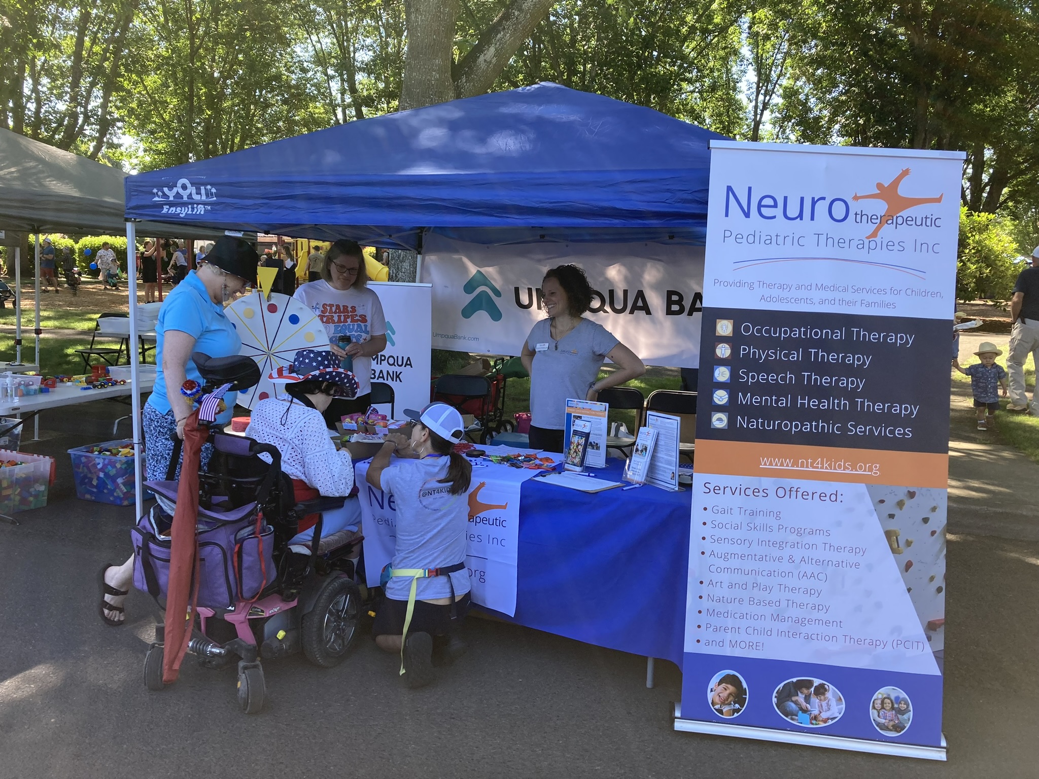 Neuro’s table at a community event outside with trees in the background. Our Executive Director is standing behind the table with her hands on her hips, smiling at community members visiting our table. One of the community members is in a wheelchair as they sign one of our clipboards. Another community member is crouched down next to them, facing them at eye-level. 