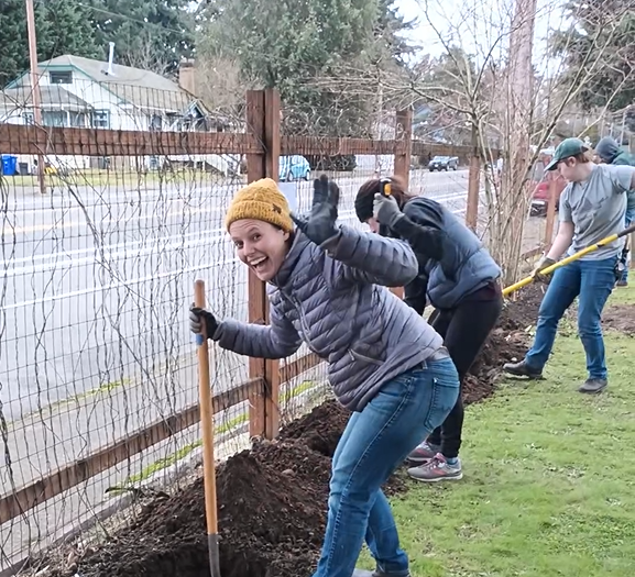 Our Executive Director, Amanda Watters, outside helping our NE Portland Clinic with their front yard revitalization. She is dressed in a winter hat and coat, a shovel in one hand as she smiles at the camera and waves as she digs.