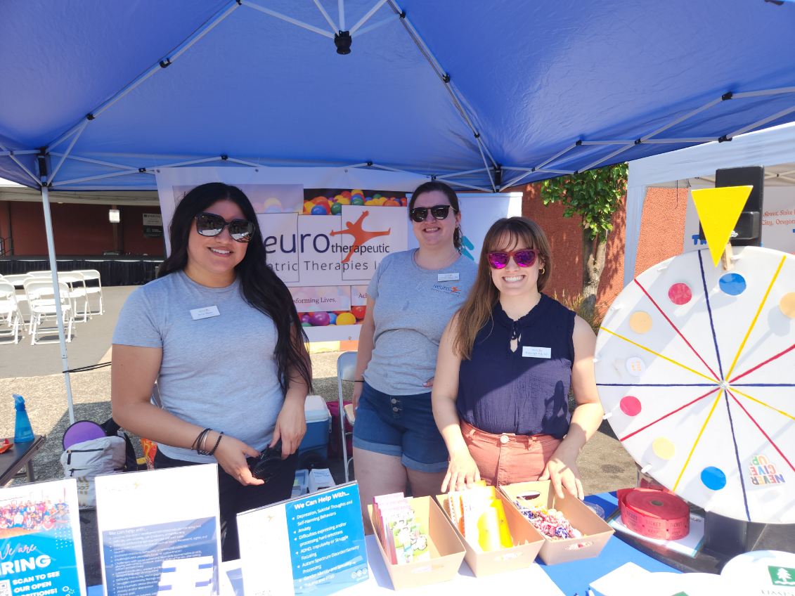 Three Neuro staff members at our Neuro table at one of our community events. The weather is bright and the sun is shining on them as all three wear shades and smile at the camera.